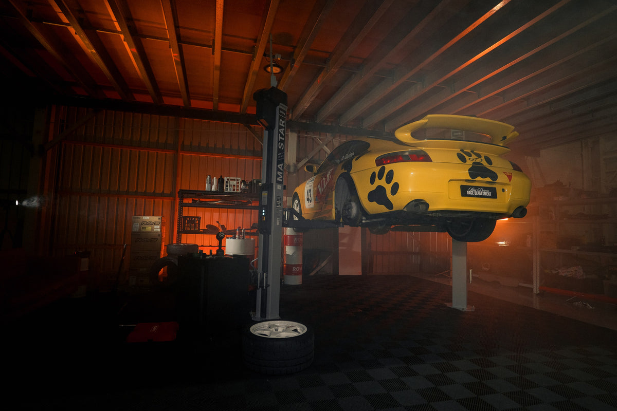 Stay Driven Race Department workshop at night, a yellow Porsche 996 GT3 race car raised on a two post hoist under red light